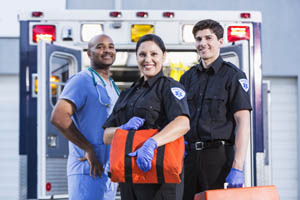 Three emergency medical professionals stand in front of an open ambulance, smiling and holding medical equipment.