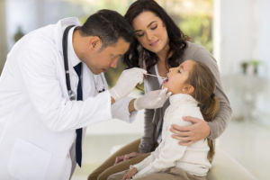 Doctor examining young girl’s throat with mother comforting her during a medical check-up in a bright, modern clinic.