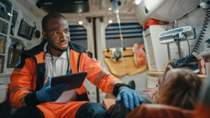 Paramedic in orange uniform holding a tablet, talking to a patient inside an ambulance, surrounded by medical equipment.