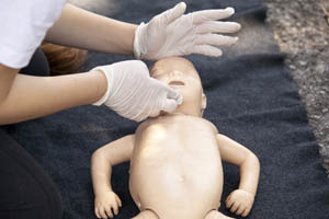 Person wearing gloves practicing infant CPR on a baby mannequin placed on a dark mat outdoors.