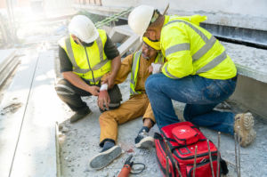Two construction workers assist an injured colleague sitting on the ground at a worksite, wearing safety gear, with a first aid kit nearby.