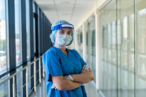 Healthcare worker in blue scrubs, face shield, mask, and gloves standing in a hospital corridor with arms crossed.