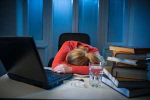 Woman with head down on desk surrounded by books, a laptop, and a glass of water, appearing exhausted or overwhelmed in a dimly lit room.