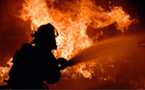 Silhouette of a firefighter spraying water to extinguish a large blaze with intense orange flames in the background.