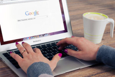Person typing on a laptop with the Google search homepage on the screen and a cup of coffee on a wooden table.