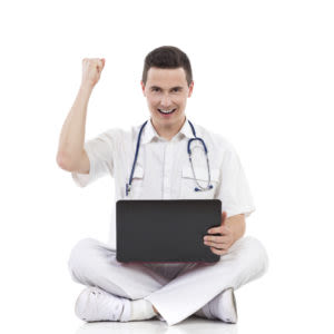 Smiling healthcare professional in white uniform, sitting cross-legged with a laptop and stethoscope, raising a fist in celebration or success against a white background.