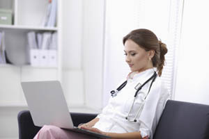 Female doctor with stethoscope sitting on a chair using a laptop in a modern medical office.