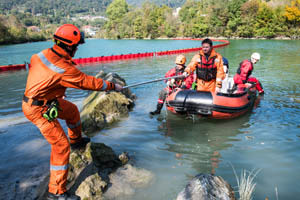 Rescue workers in orange suits pull a boat with teammates to shore on a river, surrounded by trees and floating barriers.