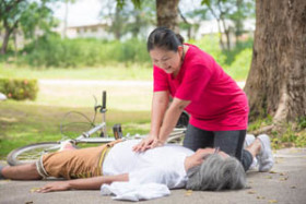 Woman performing CPR on an unconscious person lying on the ground near a bicycle in a park.