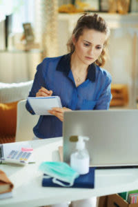 Woman in blue blouse working at a laptop, taking notes, with a mask, calculator, and hand sanitizer on the desk in a home office setting.
