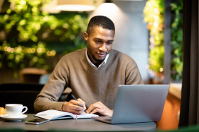 Man sitting at a table in a cozy cafe, writing in a notebook while working on a laptop, with a cup of coffee and a croissant nearby.
