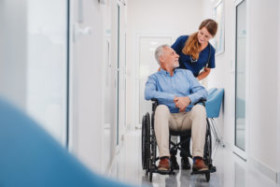 Nurse smiling and assisting an elderly man in a wheelchair down a bright hallway in a medical facility.