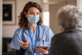 Nurse wearing a face mask and scrubs speaks with an elderly patient indoors, holding a digital tablet and gesturing with her hand.