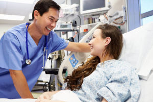 Smiling nurse talks to a female patient lying in a hospital bed in a modern medical room, both appearing happy and engaged.