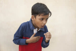Young boy in a blue and red shirt coughing with one hand on his chest, standing against a plain light-colored background.