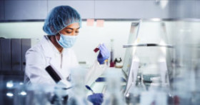 Scientist wearing a lab coat, mask, gloves, and hairnet works with test tubes and samples in a modern laboratory setting.