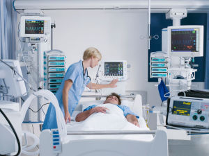 Nurse monitoring a patient in a hospital bed surrounded by medical equipment and vital sign monitors in an intensive care unit.