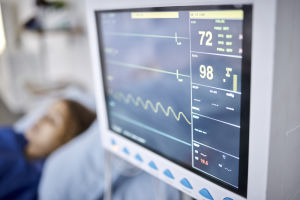Close-up of a hospital patient monitor displaying vital signs, with a blurred patient lying in bed in the background.