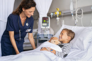Nurse smiling at a young boy lying in a hospital bed, holding a stuffed animal, with medical equipment visible in the background.