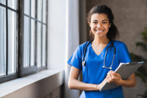 Smiling nurse in blue scrubs with a stethoscope around her neck holding a clipboard, standing by a window.