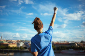 Healthcare worker in scrubs and gloves raising a fist triumphantly outdoors with a city skyline and blue sky in the background.