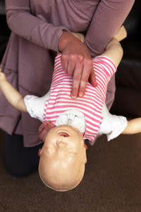 Adult demonstrating infant choking first aid on a baby manikin in a striped outfit, with the manikin held face down across the adult's knees.