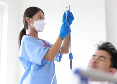 Nurse wearing mask and gloves adjusting IV drip for a patient in a hospital room.