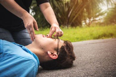 Person performing rescue breathing, pinching the nose of an unconscious man lying on the ground outdoors, preparing to give mouth-to-mouth resuscitation.