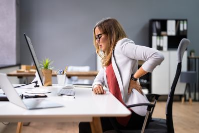 Woman sitting at a desk in an office, holding her lower back and appearing to be in pain, possibly from poor posture or prolonged sitting.